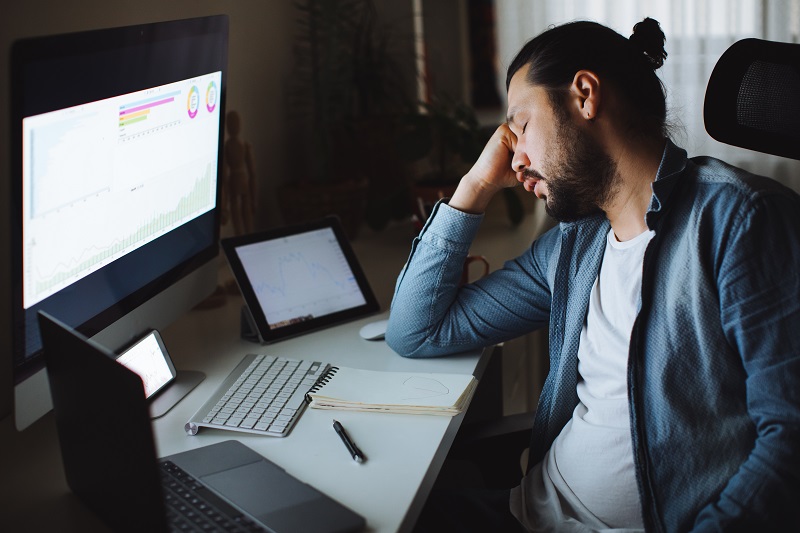 Young man closing his eyes, resting his head on his hand while sitting at his desk in front of his computer at home, struggling to stay awake. Suffering from sleep deprivation. 