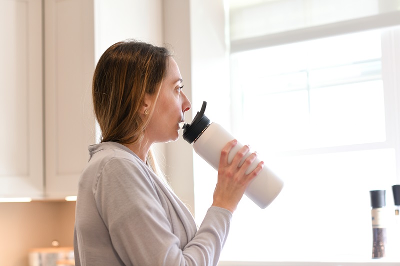 Woman drinking from a stainless steel water bottle, looking out the window. 