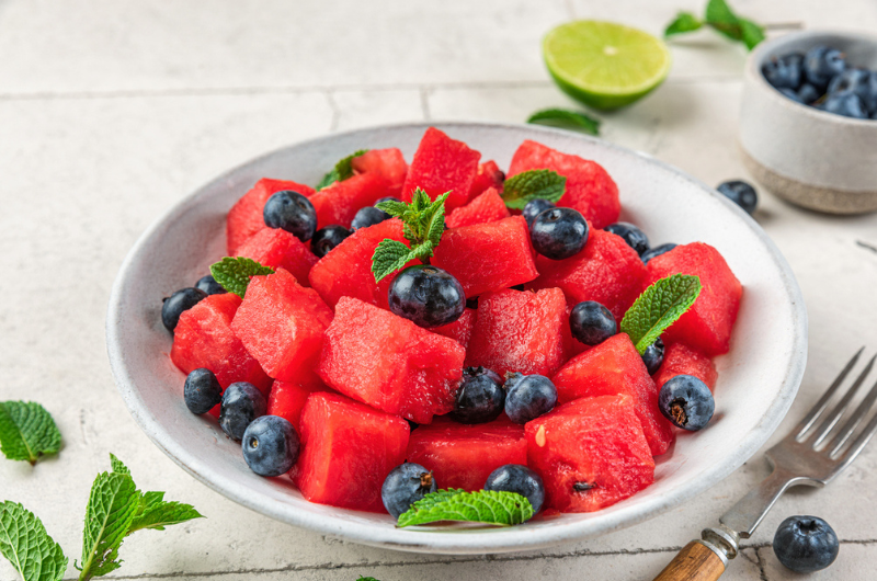 A white bowl is filled with fresh watermelon chunks and blueberries that are sprinkled with mint leaves.