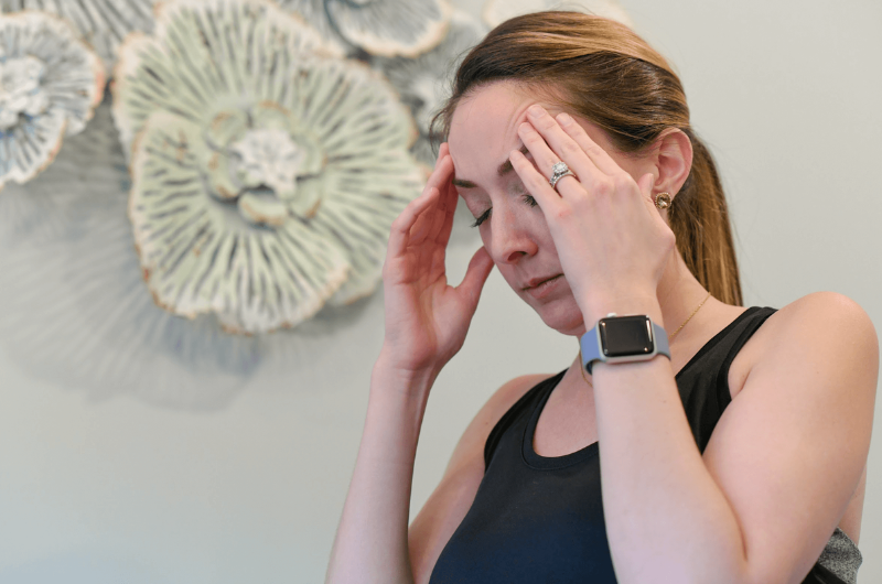 A young woman, who appears to be in pain, presses both hands to her forehead.