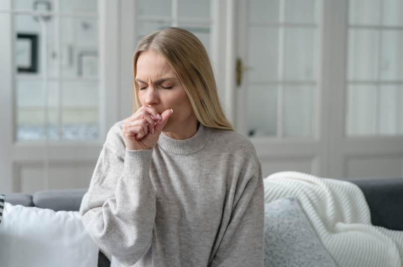 Blonde woman sits on a couch and coughs into her closed hand.