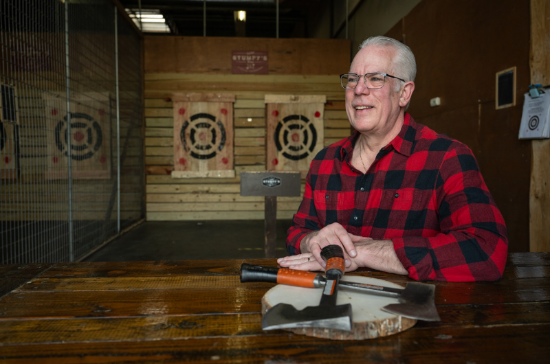 With a background of an ax-throwing lane, Scott Knox looks off camera and smiles.