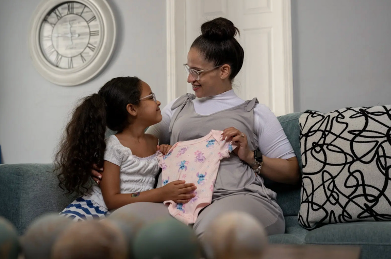 Swanny Fernandez holds a baby onesie while sitting on the couch next to a little girl.