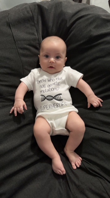 Baby Benny lying down, wearing a onesie that says "Know who else has genetic mutations? Super Heroes"